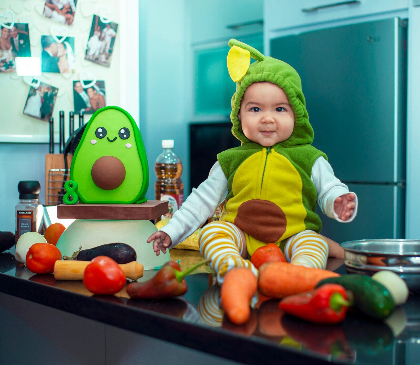 Cute baby wearing an avocado costume sitting on a kitchen counter with vegetables.